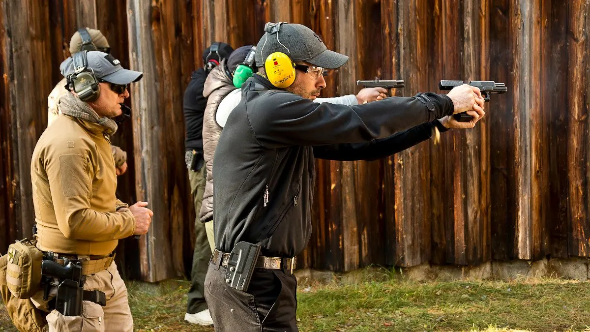 Shooters wearing ear protection fire pistols in a line by a wooden wall, watched by a man in tan clothing.